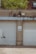 Two closed garage doors, separated by a concrete column, with a small door on the left. A sign with a warning is posted on the column. The building appears weathered with visible stains, and there are green trees and brick buildings in the background.