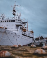 A large ship docked at a port, with intricate antennae and equipment on its deck. The vessel appears to be industrial or possibly a research boat, given its advanced and technical-looking structures. The ship is white, and there are lifeboats hanging on the sides. The background shows a cloudy sky and some structures or trees in the distance.