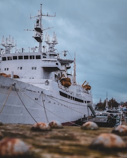 A large ship docked at a port, with intricate antennae and equipment on its deck. The vessel appears to be industrial or possibly a research boat, given its advanced and technical-looking structures. The ship is white, and there are lifeboats hanging on the sides. The background shows a cloudy sky and some structures or trees in the distance.