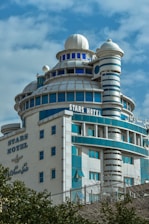 A multi-story building with a futuristic design, featuring blue and white patterns, various circular structures, and a dome at the top. The name 'Stars Hotel' is displayed prominently on the façade in both English and another language. A clear blue sky serves as the backdrop.