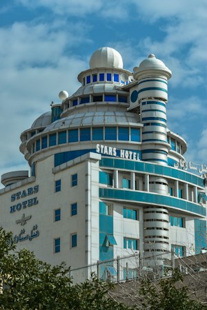 A multi-story building with a futuristic design, featuring blue and white patterns, various circular structures, and a dome at the top. The name 'Stars Hotel' is displayed prominently on the fa&ccedil;ade in both English and another language. A clear blue sky serves as the backdrop.