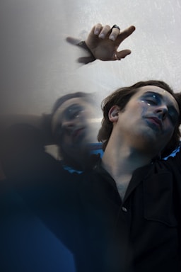 A person with dramatic makeup, featuring dark eye shadow and tears streaming down the cheek, lies against a reflective surface. The hand is raised, adorned with rings, and the image conveys a sense of introspection or melancholy, emphasized by the blurred reflection.