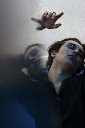 A person with dramatic makeup, featuring dark eye shadow and tears streaming down the cheek, lies against a reflective surface. The hand is raised, adorned with rings, and the image conveys a sense of introspection or melancholy, emphasized by the blurred reflection.