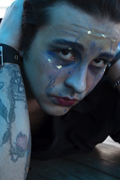 A person with artistic face paint featuring blue hues and gold accents looks intently at the camera. The individual has tattoos on the arm, visible with a detailed floral design. They are wearing a dark-colored watch and have a contemplative expression.