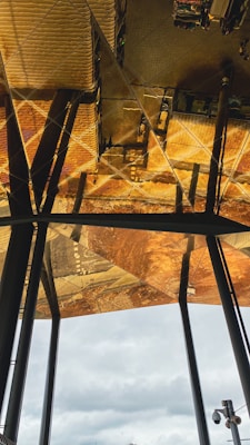 A reflective ceiling panel with a golden and bronze color palette creates an intricate pattern. Steel beams vertically intersect the reflection, and below, a cloudy sky is visible.
