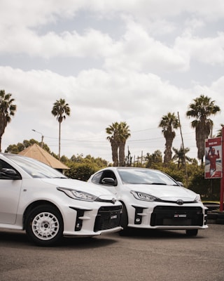 Two white cars are parked side by side on a paved area, with a background of palm trees and partly cloudy skies. A signboard with a promotional image is visible in the background.
