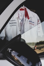 A white Toyota racing team shirt with various sponsor logos hangs on a hanger inside a car, visible through the window. The reflection of the surroundings can be seen on the side mirror of the car.