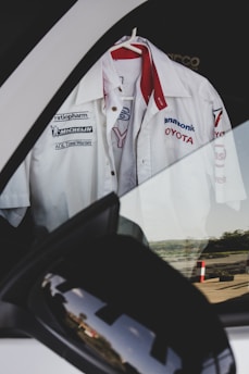 A white Toyota racing team shirt with various sponsor logos hangs on a hanger inside a car, visible through the window. The reflection of the surroundings can be seen on the side mirror of the car.