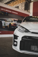 A sleek, modern white car is positioned in front of a poster featuring a racing car and the slogan 'Life's Mission: Winning.' The setting appears to be indoors with beige and red elements on the wall and a grid pattern visible above the poster.