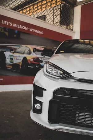 A sleek, modern white car is positioned in front of a poster featuring a racing car and the slogan 'Life's Mission: Winning.' The setting appears to be indoors with beige and red elements on the wall and a grid pattern visible above the poster.