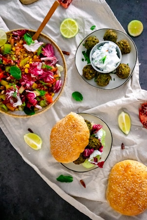 A flat lay of a vibrant, colorful salad beside a plate containing falafel balls with a creamy dipping sauce, all placed on a light-colored cloth. Surrounding these are lemon slices, leafy greens, and sesame seed burger buns with a filling of falafel and radicchio.