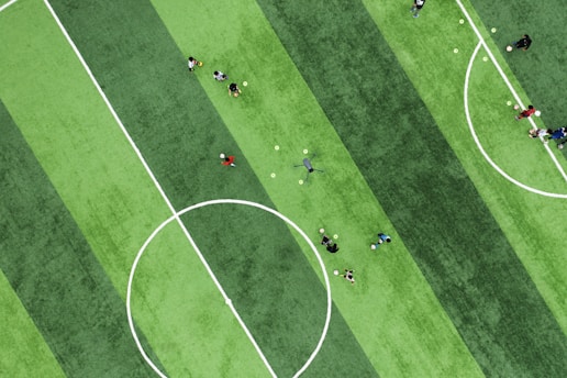 An aerial view of a soccer field with distinct green stripes and a group of people scattered around, some holding soccer balls. The central circle and white lines mark the field. The individuals appear to be engaging in a sports activity, possibly practicing or training.