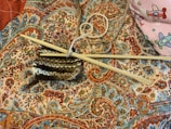 Knitting needles and a piece of partially knitted work rest on an intricately patterned fabric with vibrant paisley and floral designs. A ball of yarn is entangled with the knitting needles, and in the corner, there is a hint of a pink fabric featuring colorful airplane designs.