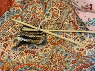 Knitting needles and a piece of partially knitted work rest on an intricately patterned fabric with vibrant paisley and floral designs. A ball of yarn is entangled with the knitting needles, and in the corner, there is a hint of a pink fabric featuring colorful airplane designs.