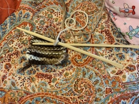 Knitting needles and a piece of partially knitted work rest on an intricately patterned fabric with vibrant paisley and floral designs. A ball of yarn is entangled with the knitting needles, and in the corner, there is a hint of a pink fabric featuring colorful airplane designs.
