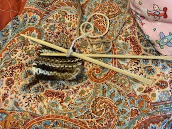 Knitting needles and a piece of partially knitted work rest on an intricately patterned fabric with vibrant paisley and floral designs. A ball of yarn is entangled with the knitting needles, and in the corner, there is a hint of a pink fabric featuring colorful airplane designs.