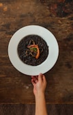 A hand holds a white plate containing a dish of dark-colored rice, possibly squid ink risotto, garnished with a piece of seafood and herbs. The background is a rustic wooden table.