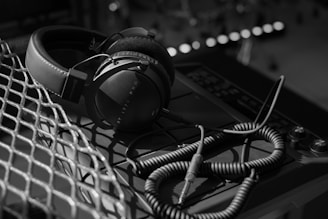 A pair of over-ear headphones with a coiled cable rests on a sound mixer. The scene is illuminated with soft lighting that creates shadows, providing a sense of depth. The headphones are positioned on a metal mesh surface, adding texture and contrast to the composition.