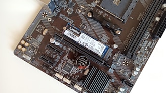 A close-up view of a computer motherboard featuring various components like a Kingston NVMe SSD, RAM slots, and a GIGABYTE logo. The board has intricate circuitry and connectors such as PCIe slots and capacitors.