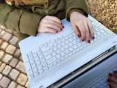 A person with red nail polish is typing on a white laptop while wearing a green jacket. The outdoor setting includes a paved surface with multi-colored cobblestones and gravel.