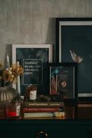 A cozy indoor scene featuring framed artwork, a stack of books, candles, and a vase with dried flowers. The framed artwork includes a quote about keeping the house green and an image of a person from behind. Books like 'Great Expectations' and 'The Master and Margarita' are visible in the stack. The atmosphere is warm and inviting.