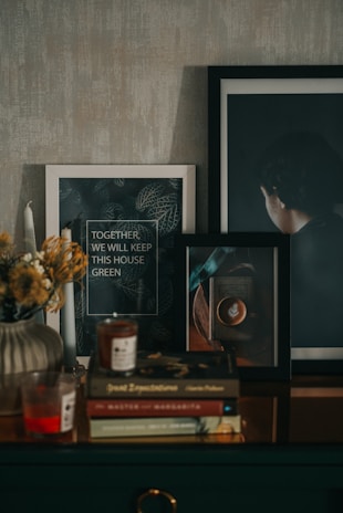 A cozy indoor scene featuring framed artwork, a stack of books, candles, and a vase with dried flowers. The framed artwork includes a quote about keeping the house green and an image of a person from behind. Books like 'Great Expectations' and 'The Master and Margarita' are visible in the stack. The atmosphere is warm and inviting.