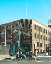 A street scene featuring a large, industrial-style brick building with a mural advertising Angel City Brewery. The street is busy with several people crossing, including a person walking a dog. The sky is clear and blue, and the area is in the Los Angeles Arts District.