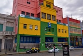 A vibrant street scene featuring a colorful building with a mix of pink, yellow, green, and blue sections. The front of the building displays the names 'Escuela Pedro de Mendoza' and 'MBQM Museo Benito Quinquela Martín.' Multiple windows and architectural details are visible. A few cars, including a black and yellow taxi, are parked on a cobblestone street. People can be seen beside and in front of the building. A bus stop with a sign is also present in the foreground.