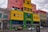 A vibrant street scene featuring a colorful building with a mix of pink, yellow, green, and blue sections. The front of the building displays the names 'Escuela Pedro de Mendoza' and 'MBQM Museo Benito Quinquela Martín.' Multiple windows and architectural details are visible. A few cars, including a black and yellow taxi, are parked on a cobblestone street. People can be seen beside and in front of the building. A bus stop with a sign is also present in the foreground.