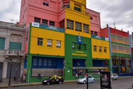 A vibrant street scene featuring a colorful building with a mix of pink, yellow, green, and blue sections. The front of the building displays the names 'Escuela Pedro de Mendoza' and 'MBQM Museo Benito Quinquela Mart&iacute;n.' Multiple windows and architectural details are visible. A few cars, including a black and yellow taxi, are parked on a cobblestone street. People can be seen beside and in front of the building. A bus stop with a sign is also present in the foreground.