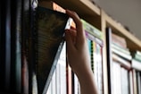 A hand is reaching for a book on a shelf filled with various books. The shelf is somewhat dimly lit, and the book being pulled out has a dark cover with some text visible.