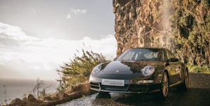 A sleek black Ferrari parked on a sunlit coastal road against a dark backdrop