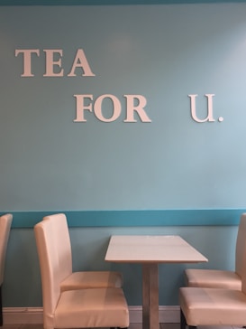 A light blue wall features white letters spelling 'TEA FOR U.' Two cream-colored upholstered chairs are positioned on either side of a small, square table with a white surface and a metallic base. The setting suggests a cozy and minimalist café or tea room environment.