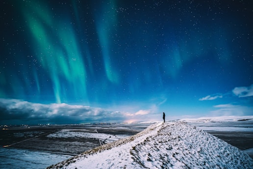A traveler looking out over a snowy landscape with a glowing aurora overhead at dawn.