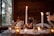 A cozy and intimate dinner setting is presented, featuring tall lit candles, a glass of red wine, and decorative autumn-themed flowers. The table is adorned with white fabric and warm, ambient candlelight creating a romantic and inviting atmosphere.