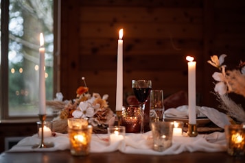 A cozy and intimate dinner setting is presented, featuring tall lit candles, a glass of red wine, and decorative autumn-themed flowers. The table is adorned with white fabric and warm, ambient candlelight creating a romantic and inviting atmosphere.