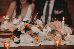 A beautifully arranged floral centerpiece featuring a variety of flowers, including white and orange blooms, with lush greenery and pampas grass. The setting is on a wooden table adorned with white fabric, surrounded by lit candles in small glass holders. In the background, there are two blurred figures, possibly a couple dressed formally.