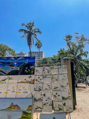 A collection of colorful paintings is displayed outdoors on a sunny day. The scenes feature vibrant landscapes, including famous landmarks and cityscapes, likely from a coastal region. A large palm tree and other lush greenery frame the background, while a clear blue sky stretches above.