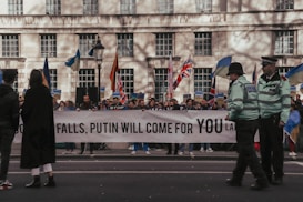 A group of people is gathered on a city street holding a large banner with a political message. Flags of different countries are visible, and police officers are standing nearby maintaining security.