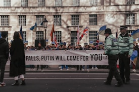 A group of people is gathered on a city street holding a large banner with a political message. Flags of different countries are visible, and police officers are standing nearby maintaining security.