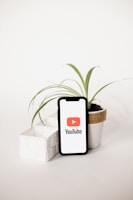 A smartphone displaying the YouTube logo stands upright on a white surface, surrounded by two small potted plants. The plant on the right has long, slender green leaves and is in a white pot wrapped with a brown band, creating a visually pleasing composition.