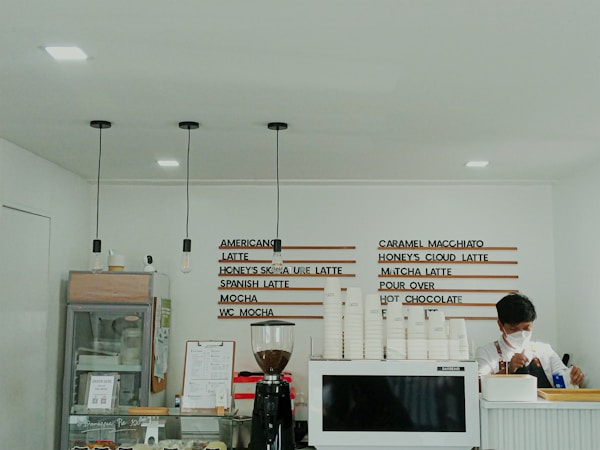 A minimalist cafe interior with a white color scheme. The menu is displayed on the wall using wooden strips with beverage options like Americano, Caramel Macchiato, and Matcha Latte. A masked barista is preparing a drink behind the counter, which features a stack of white cups, an espresso machine, and a coffee grinder. Pendant lights hang from the ceiling, and there is a glass display with desserts.