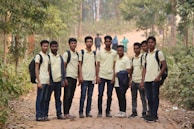 A group of young men stand on a dirt path surrounded by lush greenery. They are all wearing similar light-colored polo shirts and carrying backpacks. The setting appears to be a natural environment, possibly a park or forested area. The path leads into the distance where more people can be seen walking.