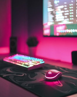 A gaming setup with a focus on a glowing RGB keyboard and a mouse on a desk. The dimly lit room has a pink hue, and a monitor displays a blurred image on the wall, adding to the immersive atmosphere.