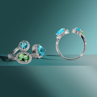 Three rings featuring oval-shaped gemstones, including a light blue stone and a green stone, set in silver bands with small accent stones. The rings are displayed on a smooth surface with a gradient turquoise background, creating a clean and elegant presentation.