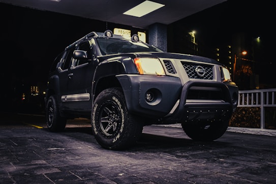 A rugged SUV is parked under a well-lit area during nighttime. The vehicle features a dark paint job, prominent front grill, and off-road tires with visible tread patterns. Some city lights and street lamps illuminate portions of the background, creating a contrast with the vehicle's body.