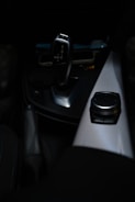 A sleek, modern car gear shift lever is prominently displayed against a dark interior background. The gear lever, which has a metallic finish, is positioned near the center console. Adjacent to the gear shift is a control knob on a textured panel, likely used for navigation or infotainment purposes.