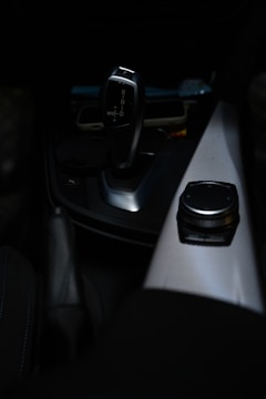 A sleek, modern car gear shift lever is prominently displayed against a dark interior background. The gear lever, which has a metallic finish, is positioned near the center console. Adjacent to the gear shift is a control knob on a textured panel, likely used for navigation or infotainment purposes.
