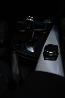 A sleek, modern car gear shift lever is prominently displayed against a dark interior background. The gear lever, which has a metallic finish, is positioned near the center console. Adjacent to the gear shift is a control knob on a textured panel, likely used for navigation or infotainment purposes.