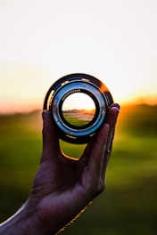A hand holding a camera lens with a landscape in focus through the lens, against a backdrop of a sunset. The sun is casting warm and vibrant light, adding a glowing effect around the lens.